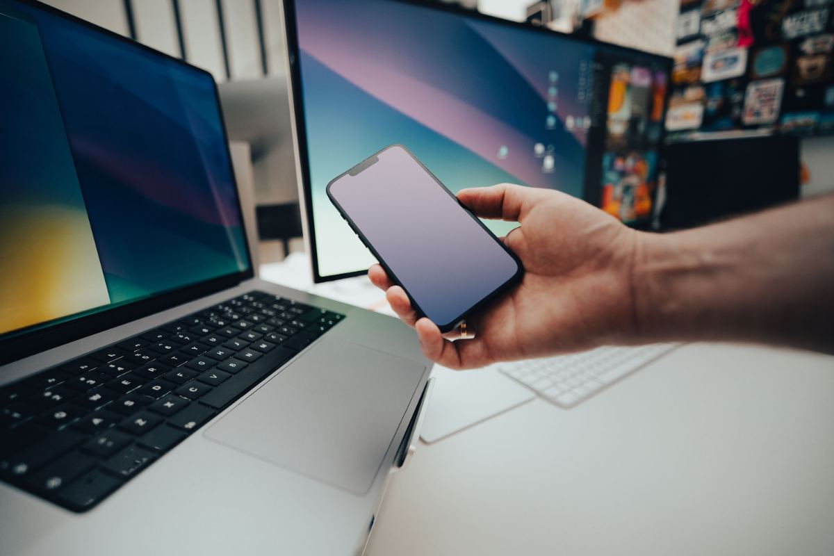 Notebook und Bildschirm an einem Arbeitsplatz, davor ein Smartphone in der Hand